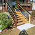 Wooden staircase leading up to a house with black railings, surrounded by greenery.